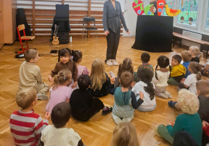Dzieci siedzą na sali gimnastycznej podczas koncertu zespołu, Pani ręce uniesione w dole.