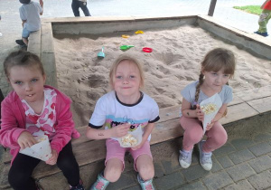Trzy dziewczynki z grupy 3 Pszczółki, siedzące w piaskownicy i jedzące popcorn