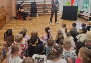Dzieci siedzą na sali gimnastycznej podczas koncertu zespołu, Pani na scenie z pluszowymi zabawkami owoców.